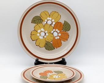 Puede incluir: Un juego de dos platos de cerámica beige con un diseño floral. Los platos presentan flores amarillas y naranjas con hojas verdes. Cada plato tiene un borde marrón. El plato más grande tiene unos 25 cm de diámetro, y el más pequeño unos 18 cm.