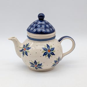 Op de afbeelding: Een crèmekleurige keramische theepot met blauwe bloemmotieven en een donkerblauw deksel versierd met witte sneeuwvlokken. De handgreep en de tuit zijn blauw. Voor het serveren van thee.