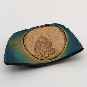 Puede incluir: Un plato de cerámica azul verdoso y marrón con un diseño de hoja. El plato está hecho a mano y tiene un aspecto rústico.