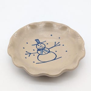 Könnte beinhalten: Ein beigefarbener Keramikteller mit gewelltem Rand, verziert mit einem blauen Schneemann-Design. Der Schneemann hat einen Zylinder, einen Schal und Arme aus Stöcken. Kleine blaue Punkte sind um den Schneemann verstreut.