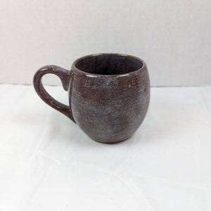 May include: A small, round, brown ceramic mug with a white glaze. The mug has a handle and a speckled appearance.
