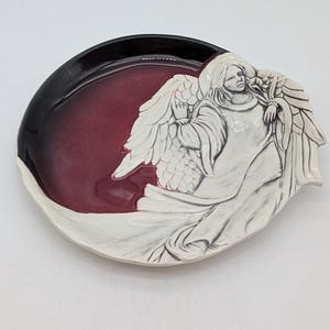 May include: Oval-shaped decorative plate with a raised relief of an angel. The plate features a white angel figure with outstretched wings against a burgundy and black background. The plate is made of ceramic.