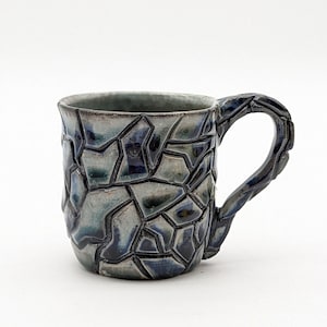 May include: A small, blue and gray ceramic mug with a geometric pattern and a handle. The mug is handmade and has a unique, textured surface.