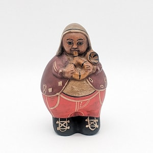 Puede incluir: Una figurita de cerámica pintada a mano de una persona que lleva una chaqueta marrón con capucha, pantalones rojos y zapatos negros. La persona sostiene un instrumento musical en sus manos.