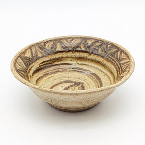 May include: A small, cream-colored ceramic bowl with a brown, geometric design around the rim. The bowl has a swirling pattern on the inside.