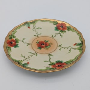 Puede incluir: Un platillo de porcelana blanca con un borde dorado y un diseño floral con flores rojas y verdes. El platillo tiene un borde festoneado y está marcado con "Bavaria" en la parte inferior.