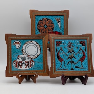 May include: Three framed ceramic tiles with Native American designs. Each tile has a turquoise background and a wooden frame. The designs include geometric patterns, figures, and pottery motifs. The tiles are displayed on small wooden stands.