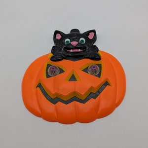 May include: Orange plastic pumpkin with a black cat peeking out of the top. The pumpkin has a black smiling face with yellow teeth and two pink eyes.