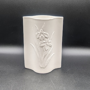 May include: White ceramic vase with an embossed design of flowers. The vase has a wavy, rectangular shape.