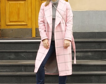 Puede incluir: Una mujer con un abrigo rosa a cuadros con cinturón a juego, un cuello alto gris y vaqueros azul oscuro. Está de pie sobre unos escalones grises, con una puerta amarilla al fondo. Lleva zapatillas blancas.