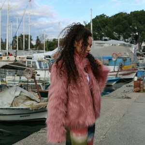 May include: A woman wearing a pink faux fur jacket and colorful tie-dye pants stands on a dock with boats in the background.