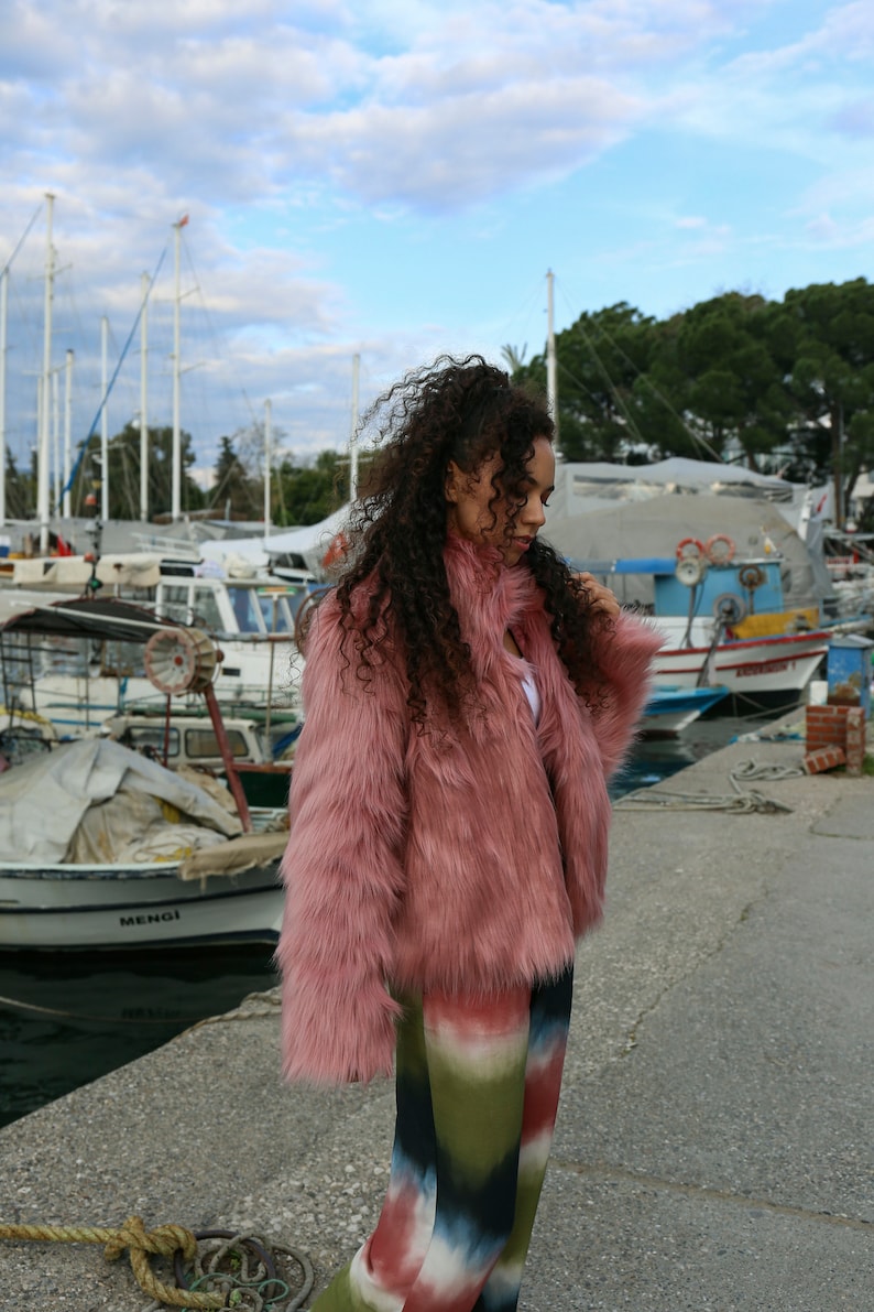 May include: A woman wearing a pink faux fur jacket and colorful tie-dye pants stands on a dock with boats in the background.