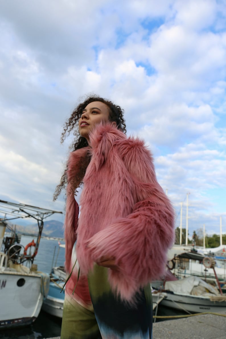 May include: A woman wearing a pink faux fur jacket with long sleeves and a fluffy texture. She is standing in front of a body of water with boats in the background.