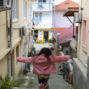 May include: A woman wearing a pink fuzzy jacket and colorful pants walks down a narrow cobblestone alleyway.  Two bicycles are parked on the right side of the alley.