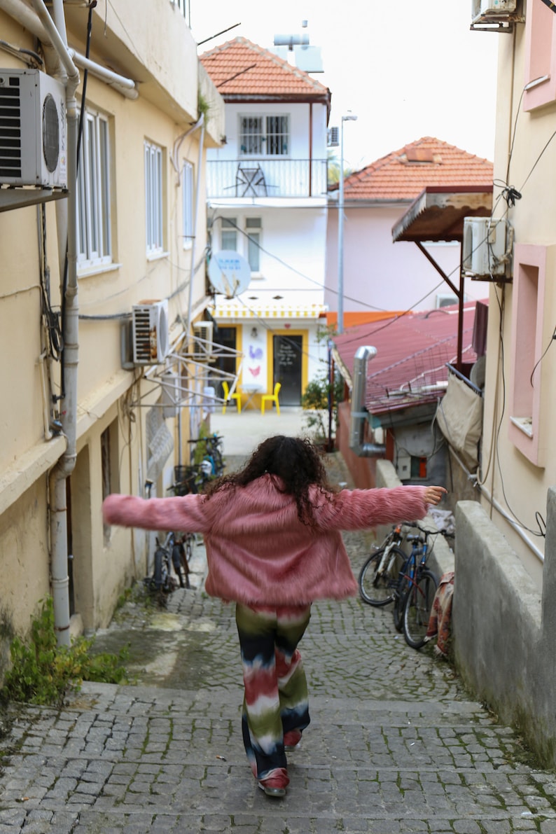 May include: A woman wearing a pink fuzzy jacket and colorful pants walks down a narrow cobblestone alleyway.  Two bicycles are parked on the right side of the alley.