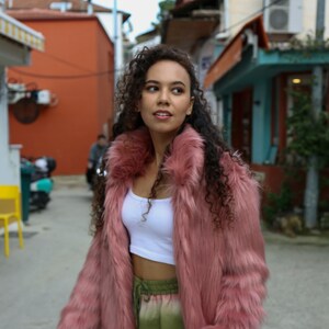 May include: A woman with curly brown hair is wearing a pink faux fur jacket, a white crop top, and green and blue tie-dye pants. She is standing in a narrow alleyway with buildings on either side.