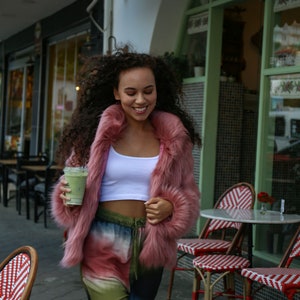 May include: A woman wearing a pink faux fur jacket, a white tank top, and tie-dye pants is holding a green smoothie. She is standing in front of a cafe with outdoor seating.
