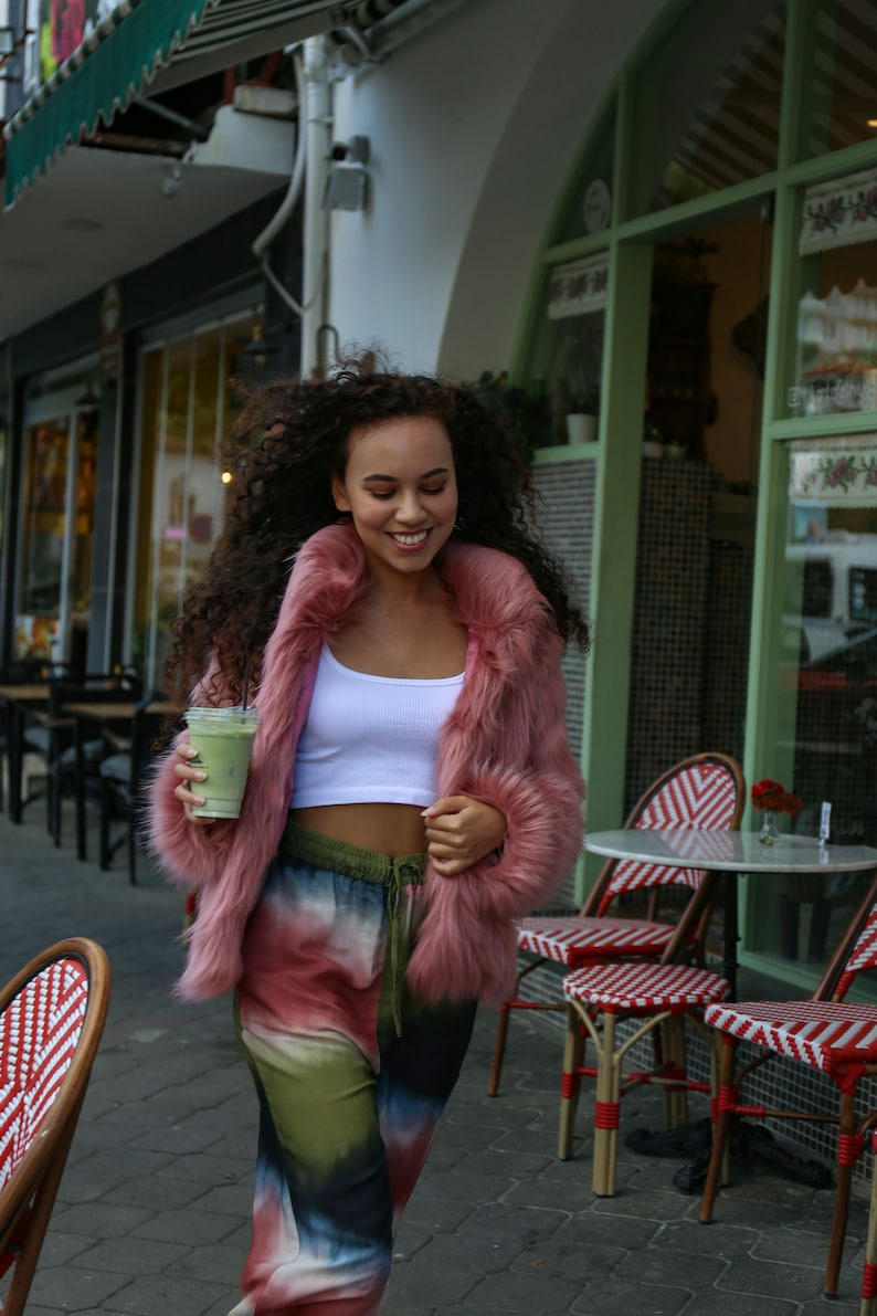 May include: A woman wearing a pink faux fur jacket, a white tank top, and tie-dye pants is holding a green smoothie. She is standing in front of a cafe with outdoor seating.