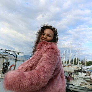 May include: A woman wearing a pink faux fur jacket with long sleeves and a fluffy texture. She is standing in front of a body of water with boats in the background. The sky is cloudy.