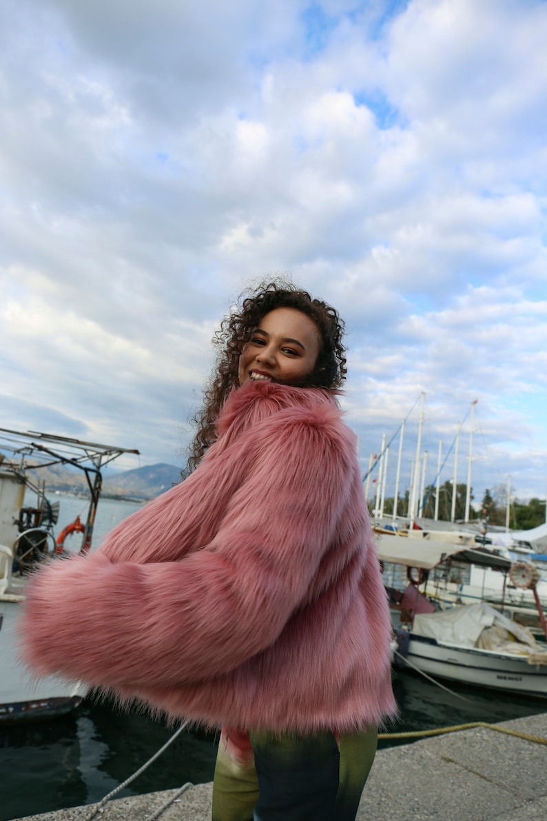 May include: A woman wearing a pink faux fur jacket with long sleeves and a fluffy texture. She is standing in front of a body of water with boats in the background. The sky is cloudy.