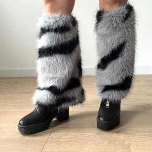 May include: A pair of faux fur leg warmers in gray and black stripes, worn over black platform boots with a zipper detail. The leg warmers have a fluffy texture and add a bold statement to the footwear.