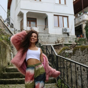 May include: A woman wearing a pink faux fur jacket, a white tank top, and tie-dye pants stands on a set of stone steps. She is smiling and looking at the camera.