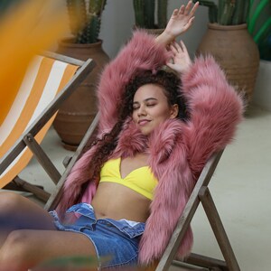 May include: A woman wearing a pink faux fur jacket, a yellow bikini top, and denim shorts is relaxing in a striped lounge chair.