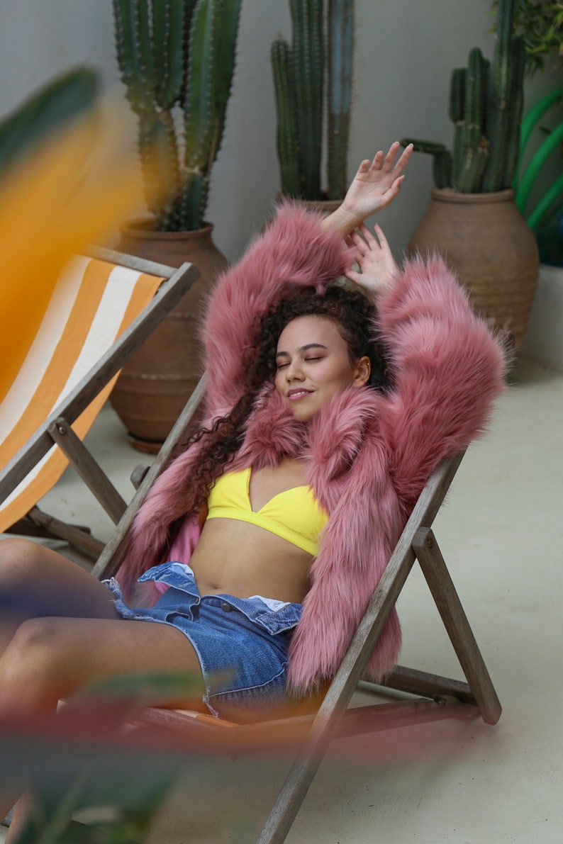 May include: A woman wearing a pink faux fur jacket, a yellow bikini top, and denim shorts is relaxing in a striped lounge chair.