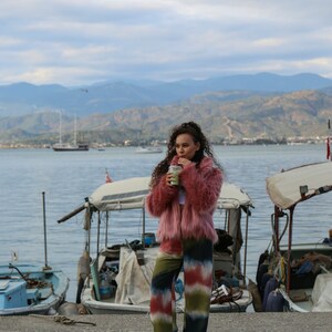 May include: A woman wearing a pink fuzzy jacket and colorful tie-dye pants stands on a dock by the water. She is holding a green beverage in her hand.