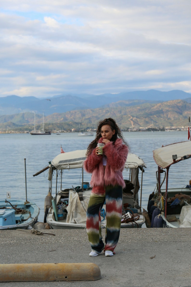 May include: A woman wearing a pink fuzzy jacket and colorful tie-dye pants stands on a dock by the water. She is holding a green beverage in her hand.