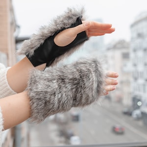 Peut inclure: Une paire de mitaines sans doigts en fausse fourrure grise avec des accents en cuir noir. Les mitaines sont portées sur les mains d'une personne.