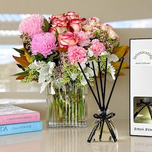 May include: A floral arrangement in a ribbed glass vase featuring pink chrysanthemums, pink-edged white roses, and green foliage. Next to it is a reed diffuser with black sticks and a box labelled Intoxicating Reed Diffuser French Pear.