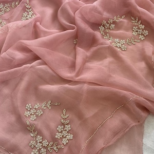 Könnte beinhalten: Ein hellrosa, durchsichtiger Saree aus Stoff mit zarter Blumen Stickerei in weißen Perlen. Der Saree hat einen dezenten Schimmer und einen weichen, fließenden Fall.