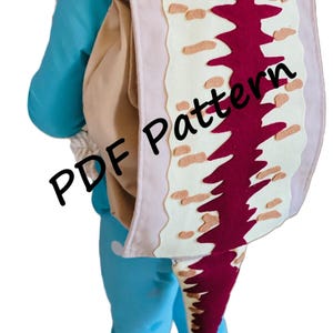 Może przedstawiać: Dziecko w niebieskim stroju z plecakiem z filcu w kształcie dinozaura. Plecak ma różowy, biały i brązowy wzór. Na plecaku widnieje napis "PDF Pattern". Pod plecakiem widnieje napis "*English Instructions".