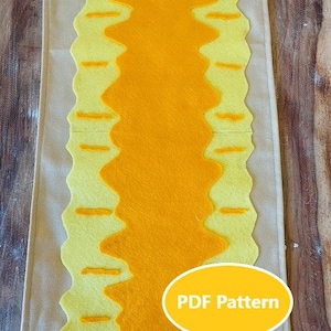 May include: A beige felt board with a yellow and orange felt design. The design is a wavy line with straight lines on either side. The board has two buttons and two white strings attached to the bottom corners. The text "PDF Pattern" is printed in yellow on a yellow oval.