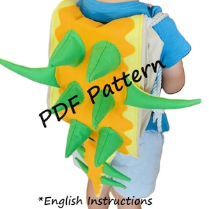 Może przedstawiać: Zielono-żółty plecak w kształcie dinozaura z ogonem i kolcami. Plecak ma biały sznurek i jest noszony przez dziecko. Na plecaku widnieje napis "PDF Pattern". Pod plecakiem widnieje napis "*English Instructions".