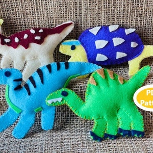 Puede incluir: Un conjunto de cinco juguetes de dinosaurio de fieltro, que incluyen un dinosaurio verde, un dinosaurio azul, un dinosaurio blanco con manchas rojas, un dinosaurio amarillo con un caparazón azul y un dinosaurio amarillo con un caparazón morado. Los juguetes están sobre un fondo de arpillera marrón. Patrón PDF.