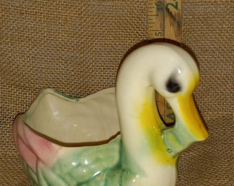 May include: A ceramic planter shaped like a white duck with yellow and green accents. The duck has a pink flower on its side and a small pot for planting.