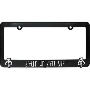 May include: Black license plate frame with the text "THIS IS THE WAY" in white, along with two Mandalorian skull emblems. The frame has a rectangular shape with rounded corners and mounting holes.
