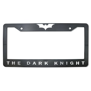 May include: Black license plate frame featuring the Batman logo at the top and the words "THE DARK KNIGHT" at the bottom in silver lettering. The frame has a rectangular shape with rounded corners and two mounting holes.