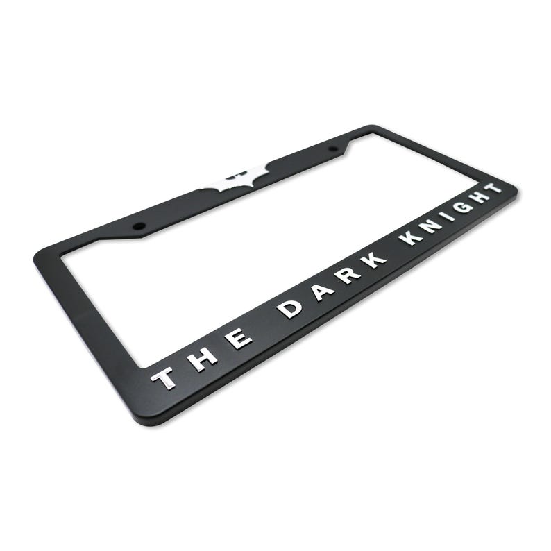 May include: Black license plate frame with the words "THE DARK KNIGHT" in white lettering. The Batman logo is visible in white on the top left corner. The frame is rectangular and has pre-drilled holes for mounting.
