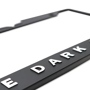 May include: Black license plate frame with the text "THE DARK KNIGHT" in raised white lettering. The frame has a sleek, modern design and is made of a durable material. Perfect for car enthusiasts.