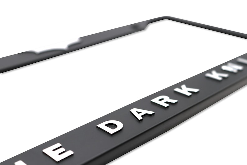 May include: Black license plate frame with the text "THE DARK KNIGHT" in raised white lettering. The frame has a sleek, modern design and is made of a durable material. Perfect for car enthusiasts.