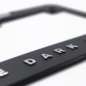 May include: Black license plate frame with raised silver lettering that reads "THE DARK KNIGHT". The frame is rectangular with rounded corners and a glossy finish. The background is white.