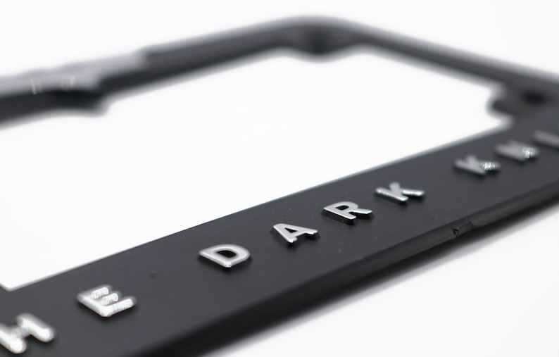 May include: Black license plate frame with raised silver lettering that reads "THE DARK KNIGHT". The frame is rectangular with rounded corners and a glossy finish. The background is white.
