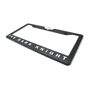 May include: Black license plate frame with the text "THE DARK KNIGHT" in white lettering. The Batman logo is at the top. The frame is rectangular and has mounting holes.