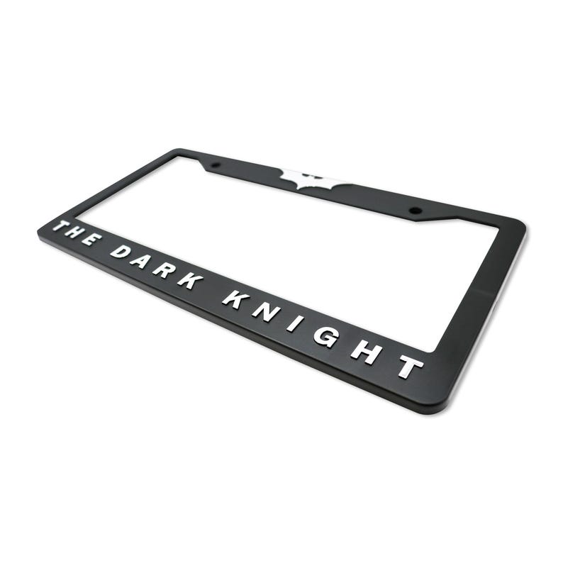 May include: Black license plate frame with the text "THE DARK KNIGHT" in white lettering. The Batman logo is at the top. The frame is rectangular and has mounting holes.