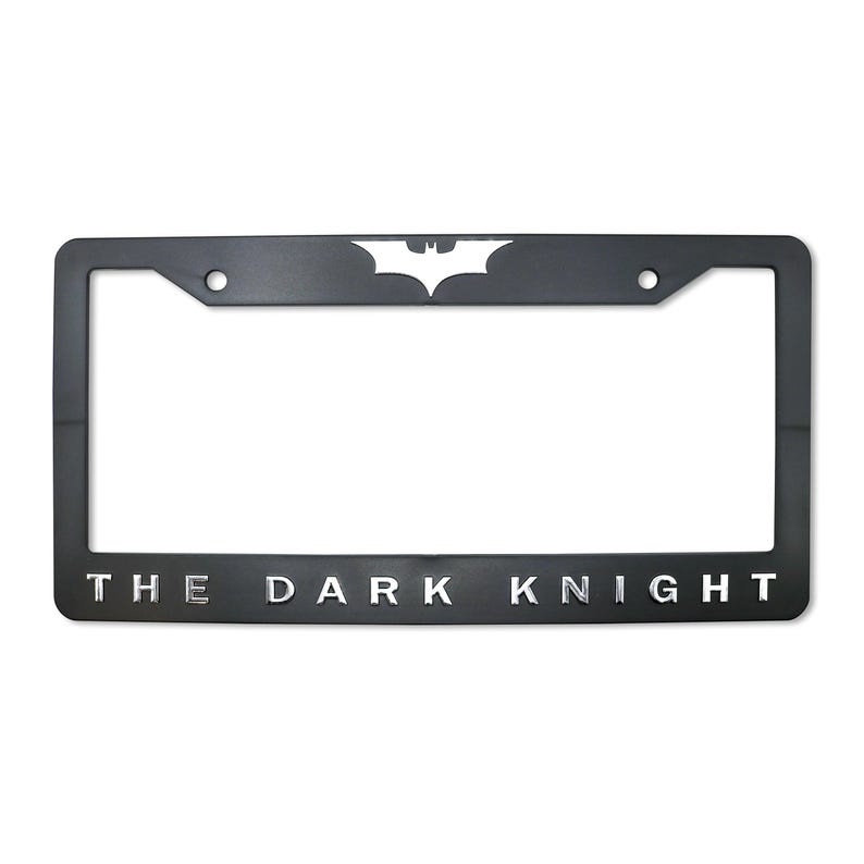 May include: Black license plate frame featuring the Batman logo at the top and the words "THE DARK KNIGHT" at the bottom in silver lettering. The frame has a rectangular shape with rounded corners and two mounting holes.