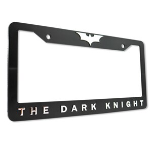 May include: Black license plate frame with the words "THE DARK KNIGHT" in silver lettering. The Batman logo is at the top center. The frame has a rectangular shape with rounded corners.