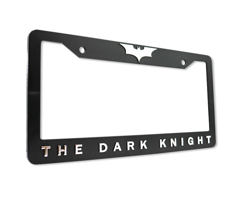 May include: Black license plate frame with the words "THE DARK KNIGHT" in silver lettering. The Batman logo is at the top center. The frame has a rectangular shape with rounded corners.